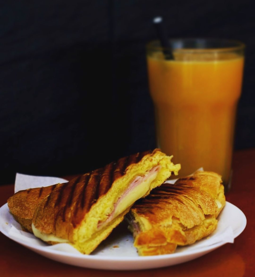 Croissant grelhado cortado ao meio, com presunto e queijo, num prato branco, ao lado de um copo de sumo de laranja