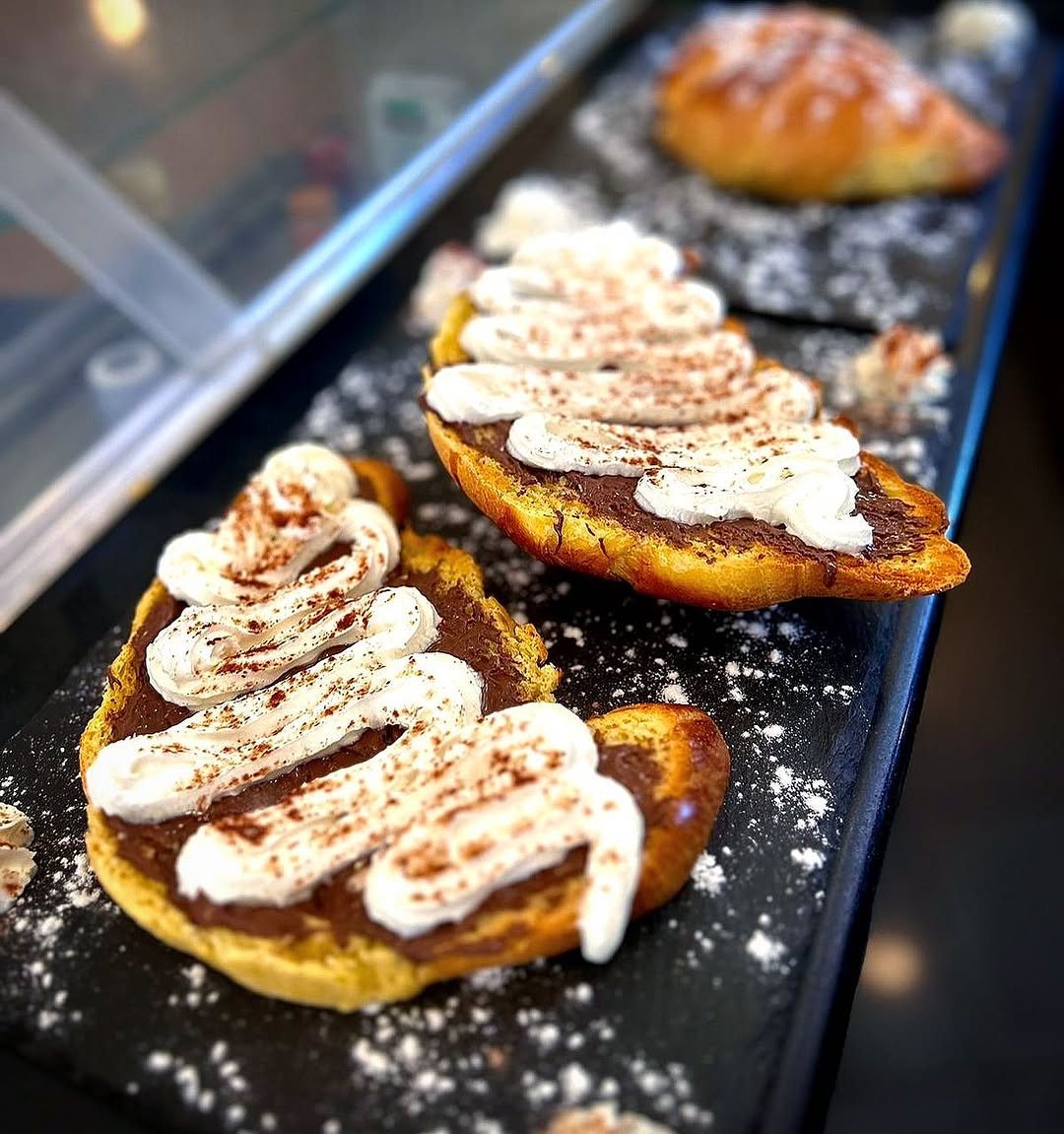 Metades de croissant cobertas com creme escuro, natas e cacau em pó, numa travessa escura com açúcar em pó